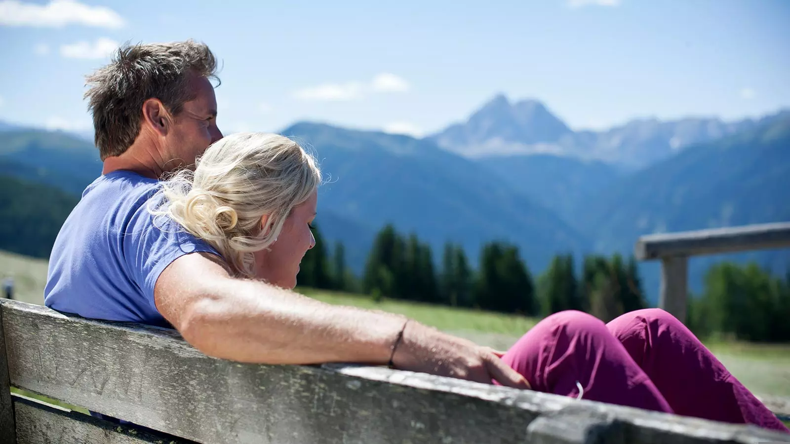 a couple is sitting on a wooden bench and enjoying the wonderful panorama of Maranza and Val Pusteria